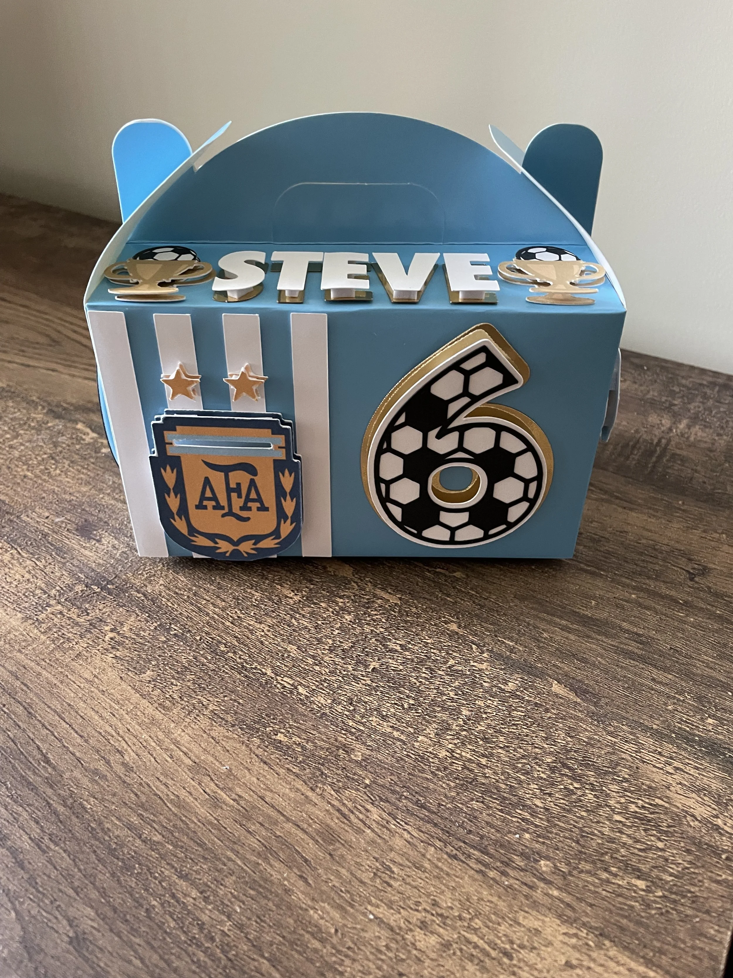 Blue and white cardboard party favor box with the name 'Steve' and a number 6, decorated with soccer and coffee cup graphics.