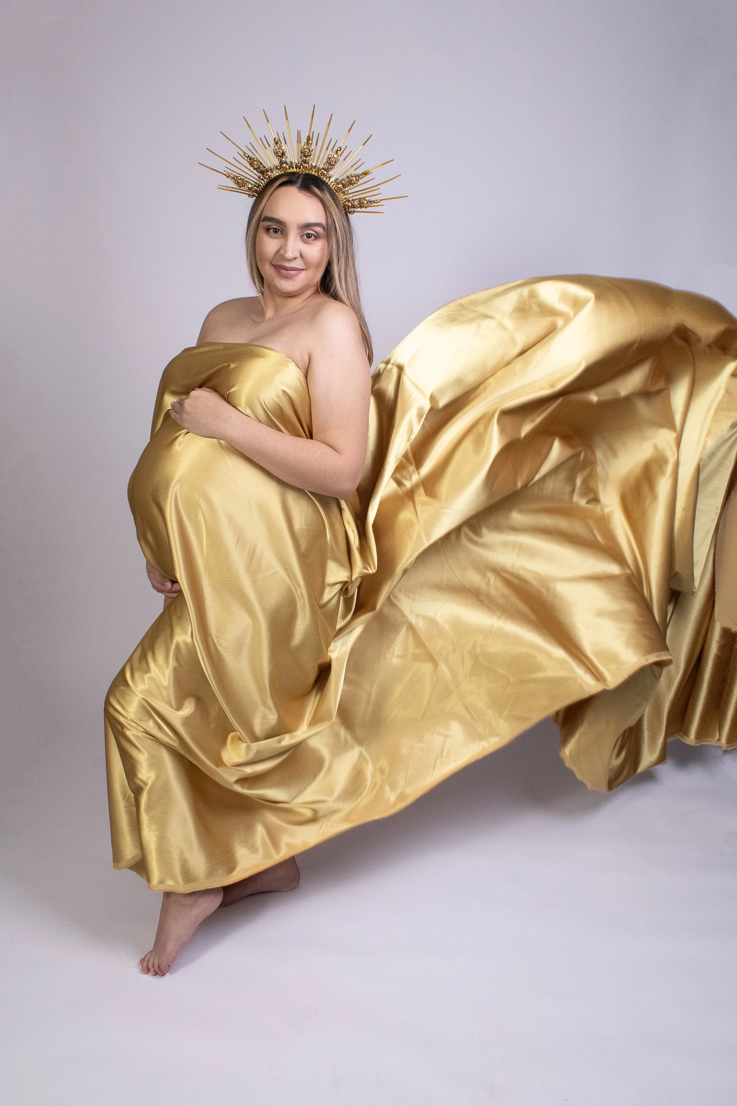 Pregnant woman in gold satin dress with a gold halo headpiece, standing on one foot on a plain light gray background.