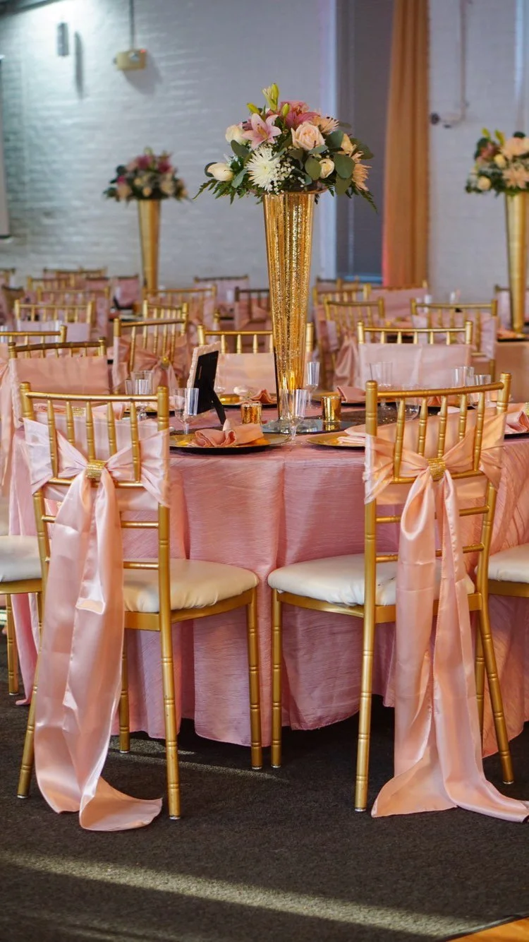 Elegant banquet table decorated with pink tablecloths, gold chairs with pink ribbons, and tall gold vases with floral centerpieces featuring pink and white flowers.