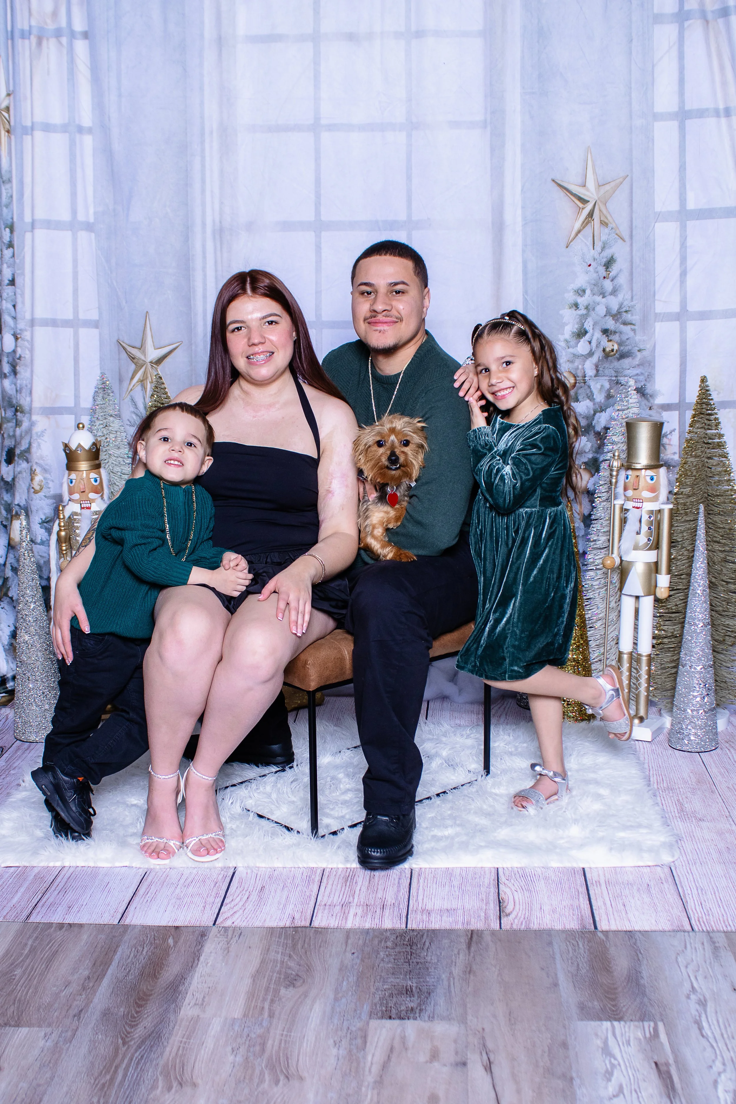 Family Christmas portrait with parents, two children, and dog, in front of holiday decorations including a white Christmas tree, nutcracker figures, and stars.