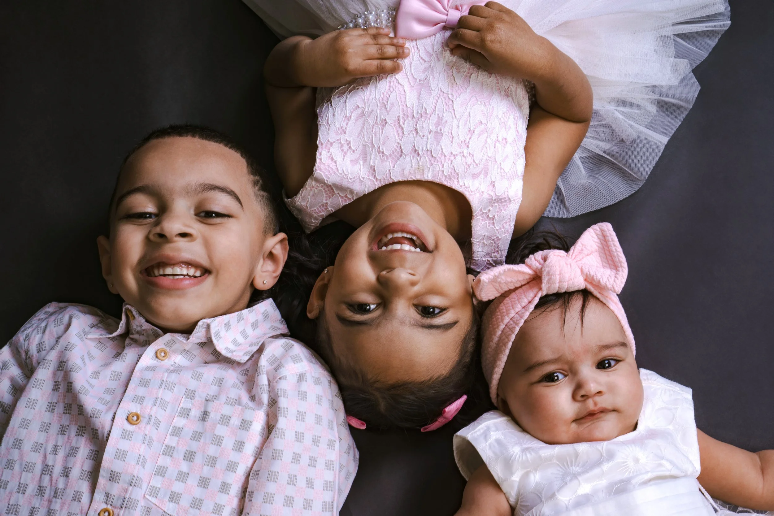 Three young girls lying on their backs, smiling and facing the camera, with a dark background.
