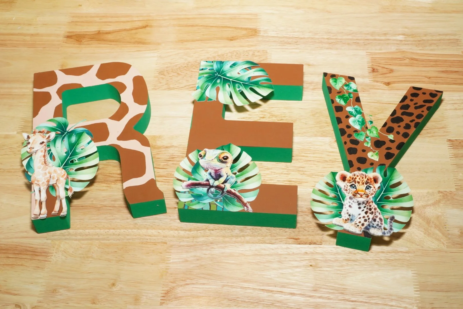 Decorative jungle-themed letters spelling 'R E Y' on a wooden surface, each letter adorned with paper animal cutouts and tropical leaves.