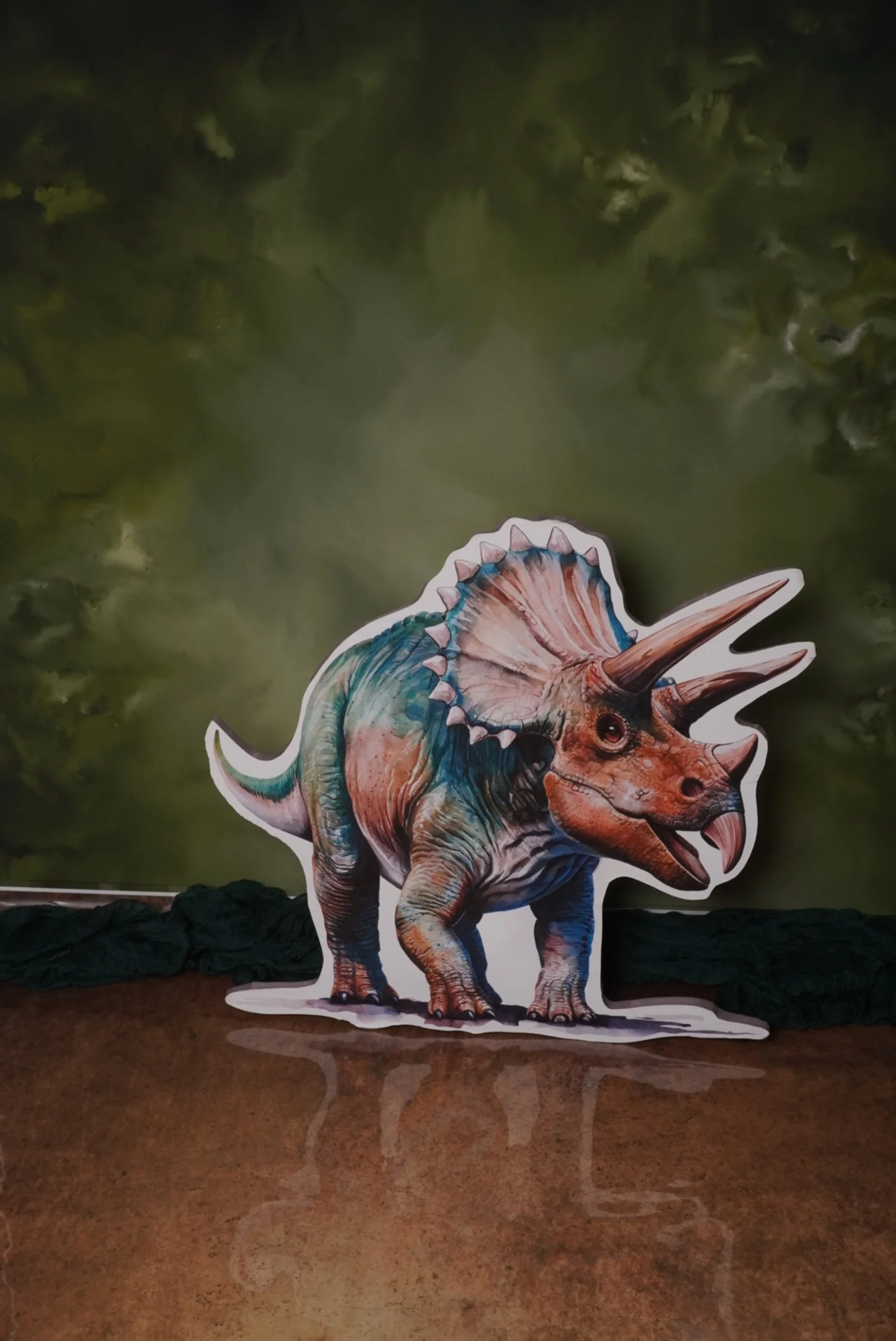 A colorful cartoon cutout of a prehistoric triceratops dinosaur on a reflective surface, with a green cloudy background.