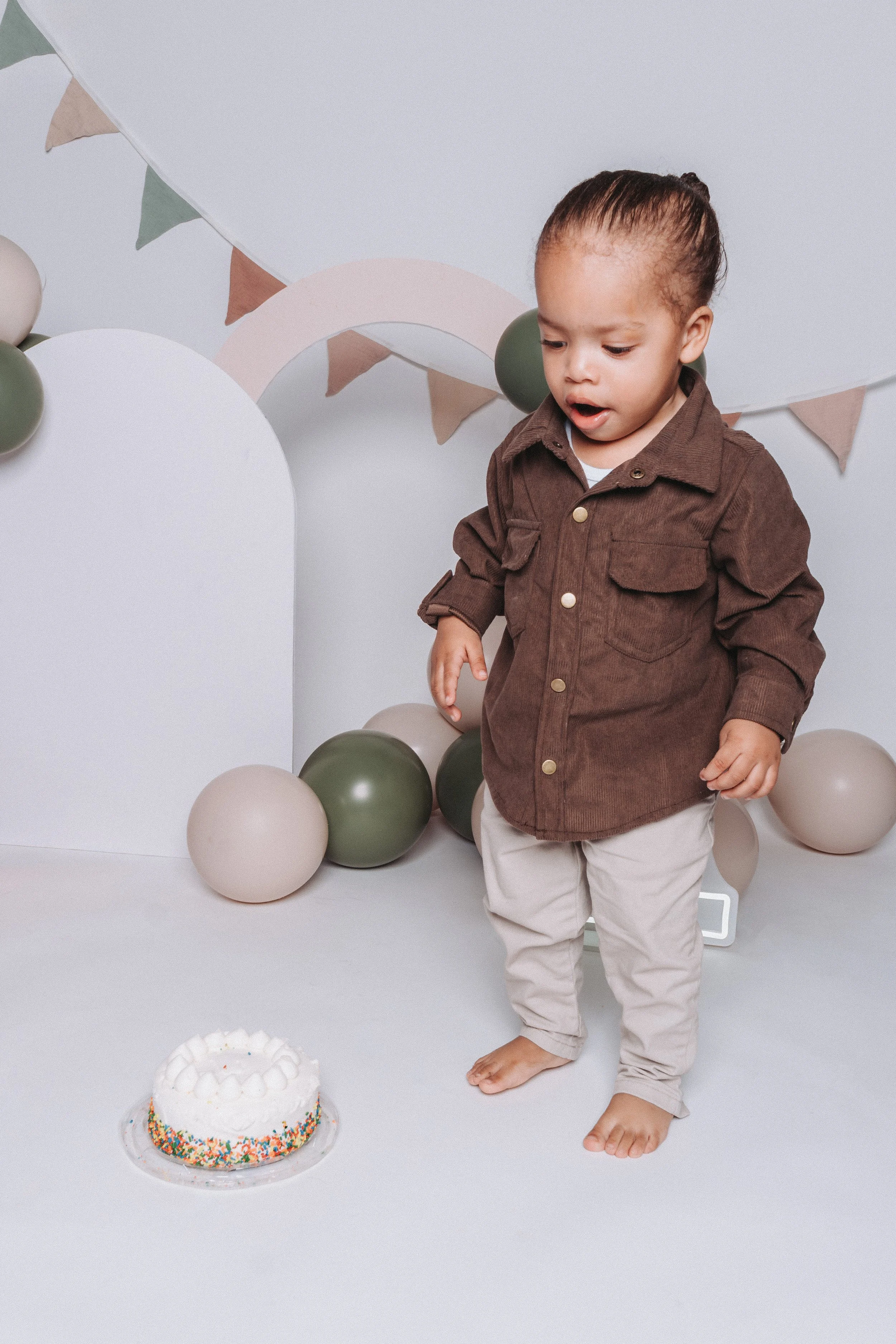 A young child wearing a brown jacket and beige pants standing in front of a birthday cake with colorful sprinkles, surrounded by neutral-colored balloons and pastel-colored decorations.