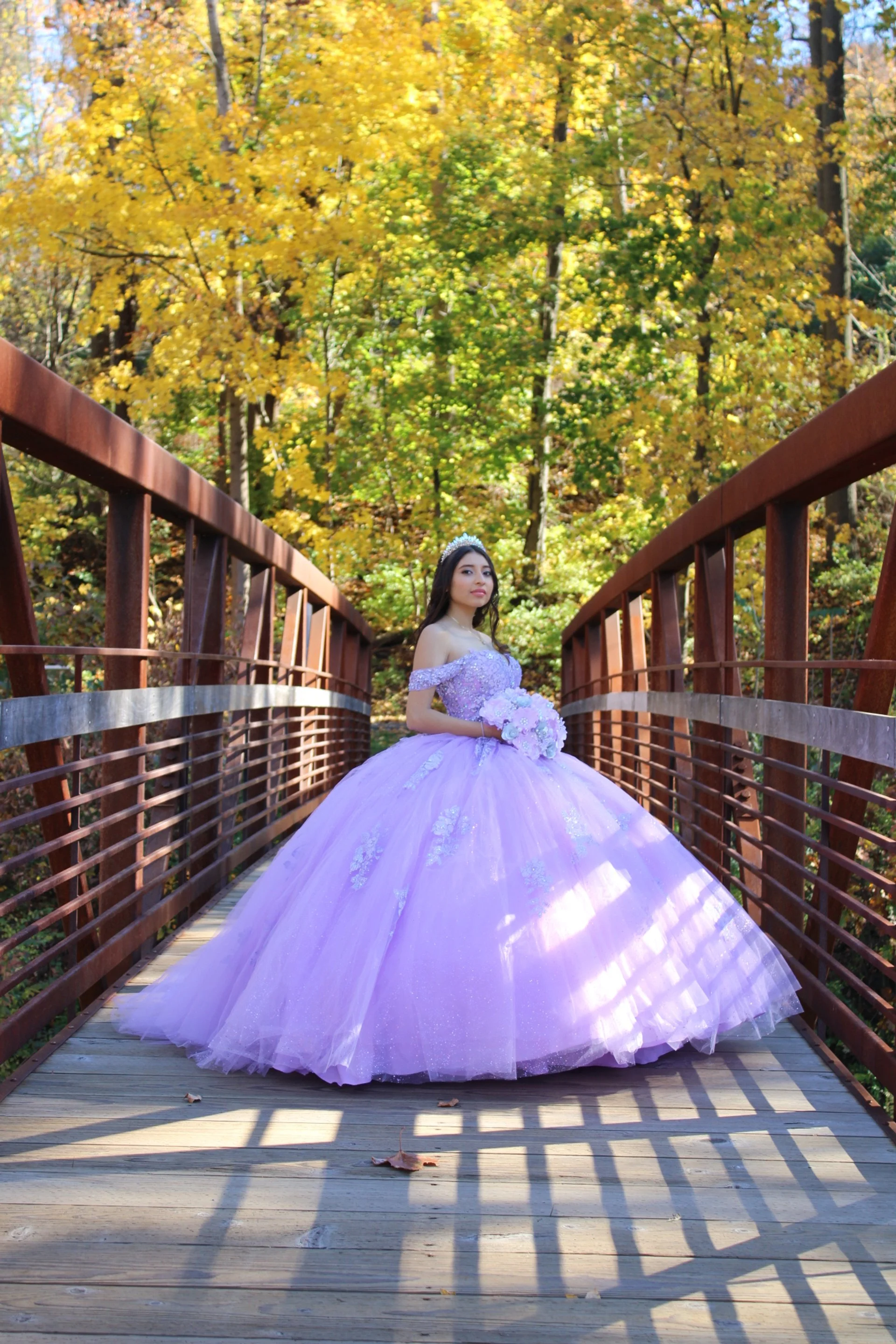 A young woman in a purple quinceañera dress holding a bouquet standing on a wooden bridge surrounded by fall foliage.