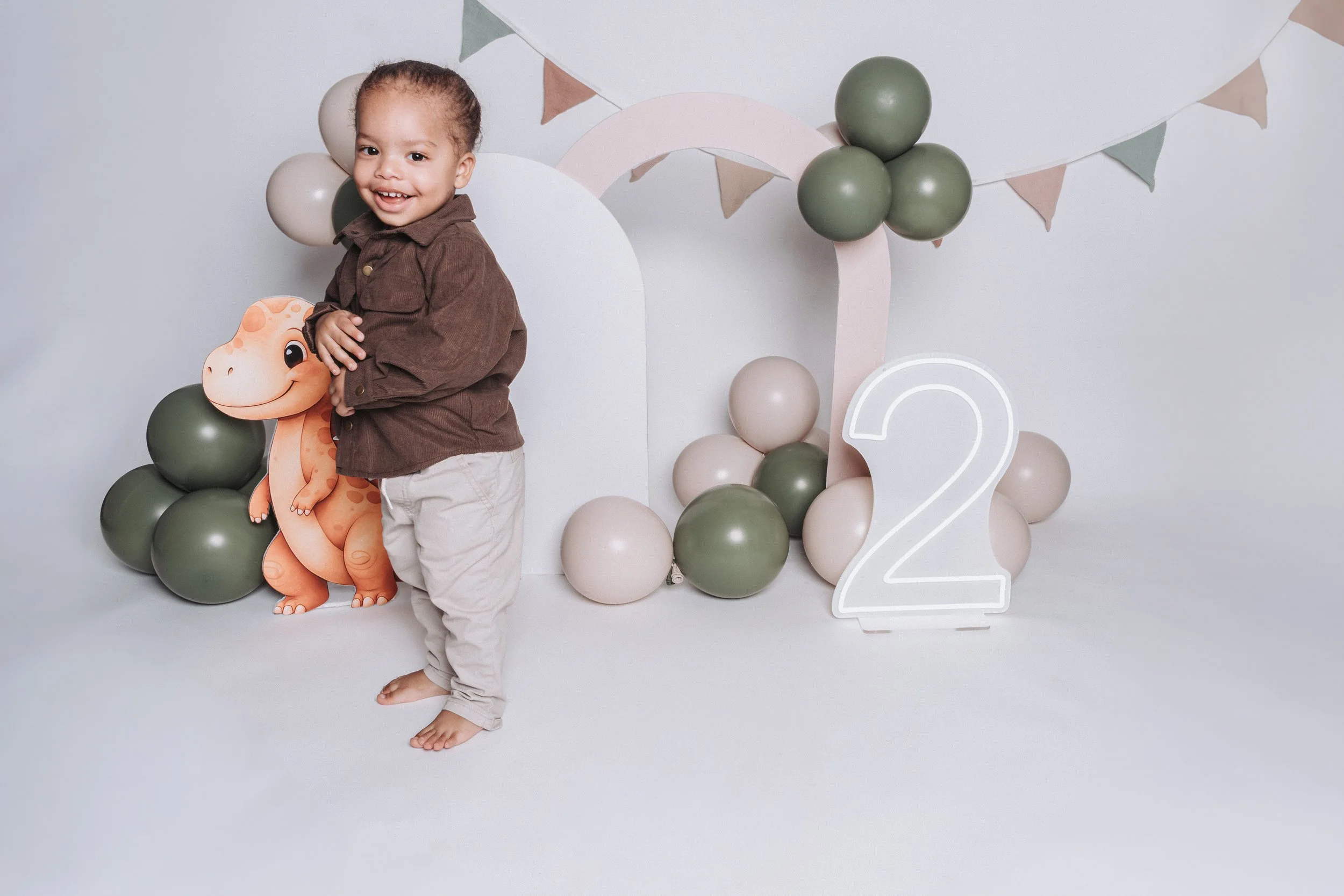A young child standing barefoot in front of a birthday celebration backdrop with balloons, a cartoon dinosaur cutout, a large white number two, and pastel-colored bunting.