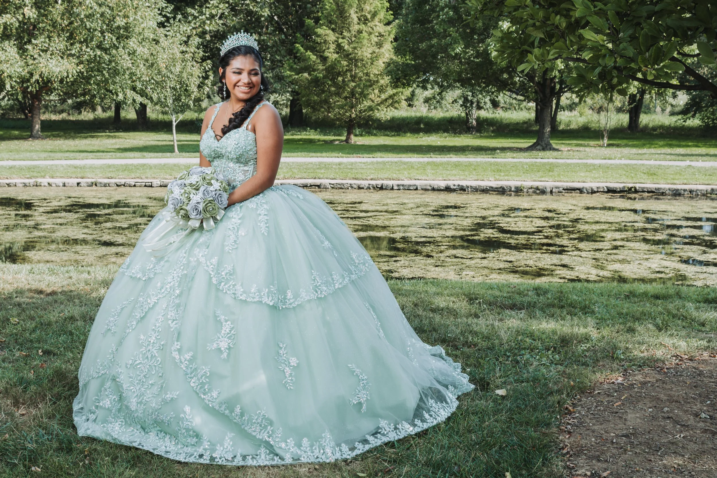 Young woman in a light blue, ornate wedding dress and tiara standing by a pond in a park, holding a bouquet of flowers, smiling happily.