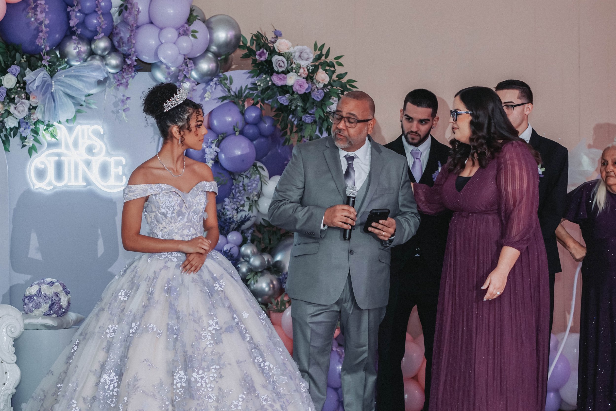 A quinceañera dressed in a lavender and white ball gown standing next to a man in a gray suit holding a microphone and a smartphone, with three people nearby at a decorated celebration with purple, white, and silver balloons and flowers.