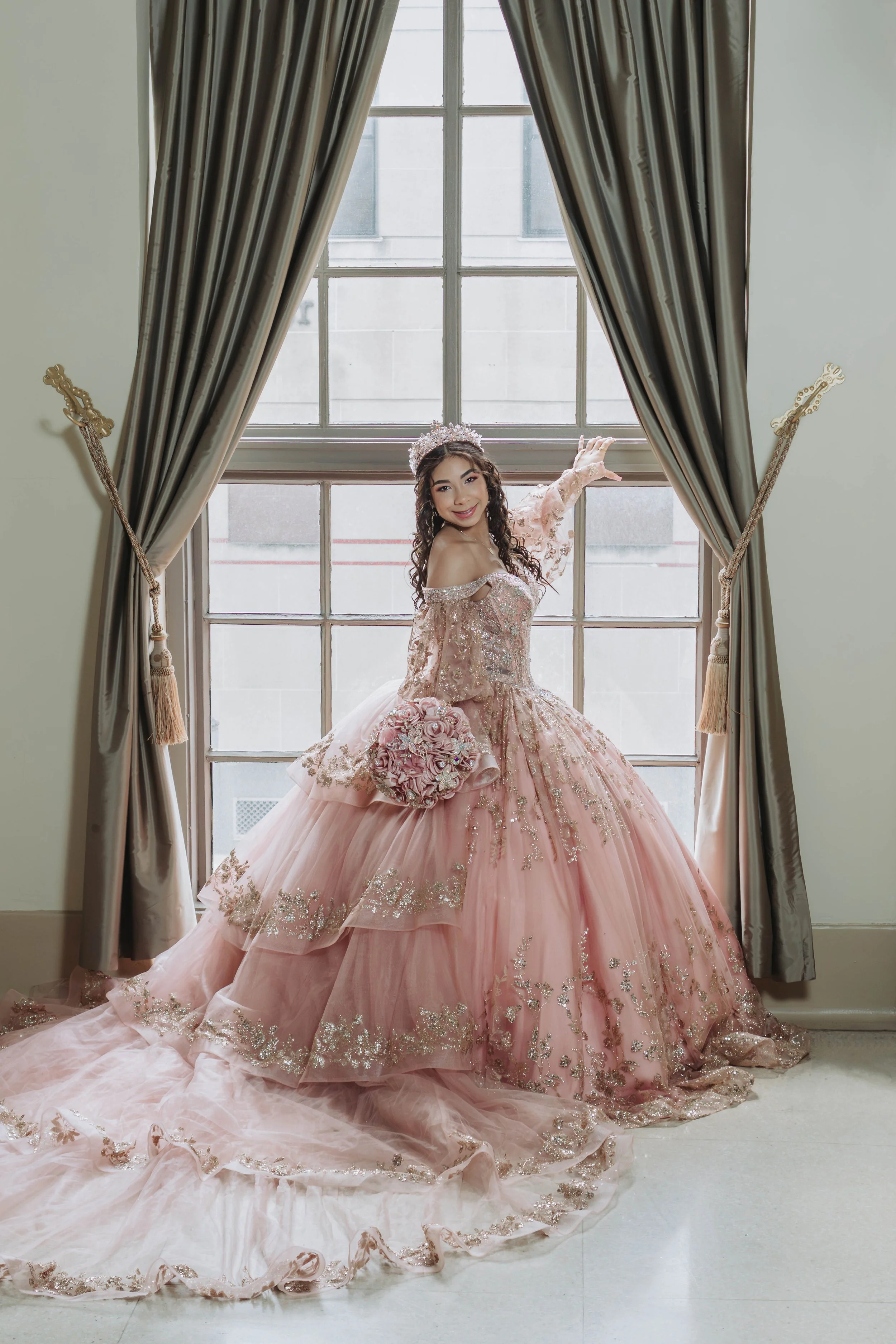 A young woman in a pink, ornate ball gown with floral details and a matching tiara, standing by a large window with gray curtains, smiling and posing.