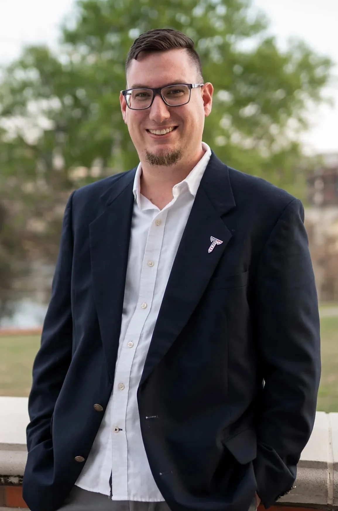 A man in glasses and a navy blazer over a white shirt, standing outdoors with blurred greenery in the background.