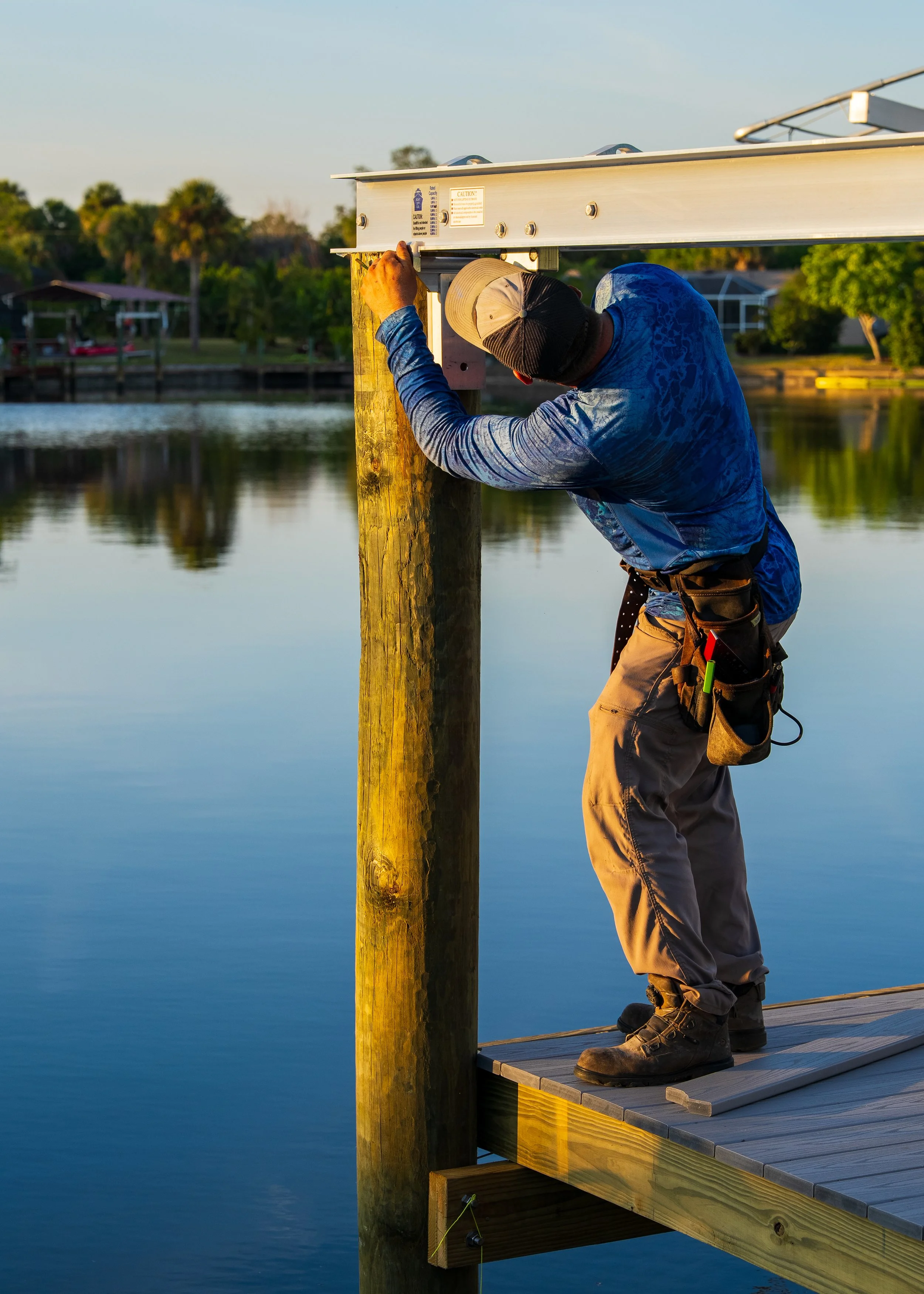 Wooden dock extending over water, surrounded by green bushes and trees, with houses and palm trees visible in the background.