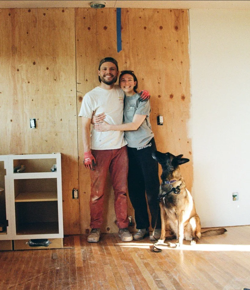 Two people, a man and a woman, standing close together, smiling, with a German Shepherd dog sitting beside them. The background shows a wooden wall with some construction or renovation work in progress.