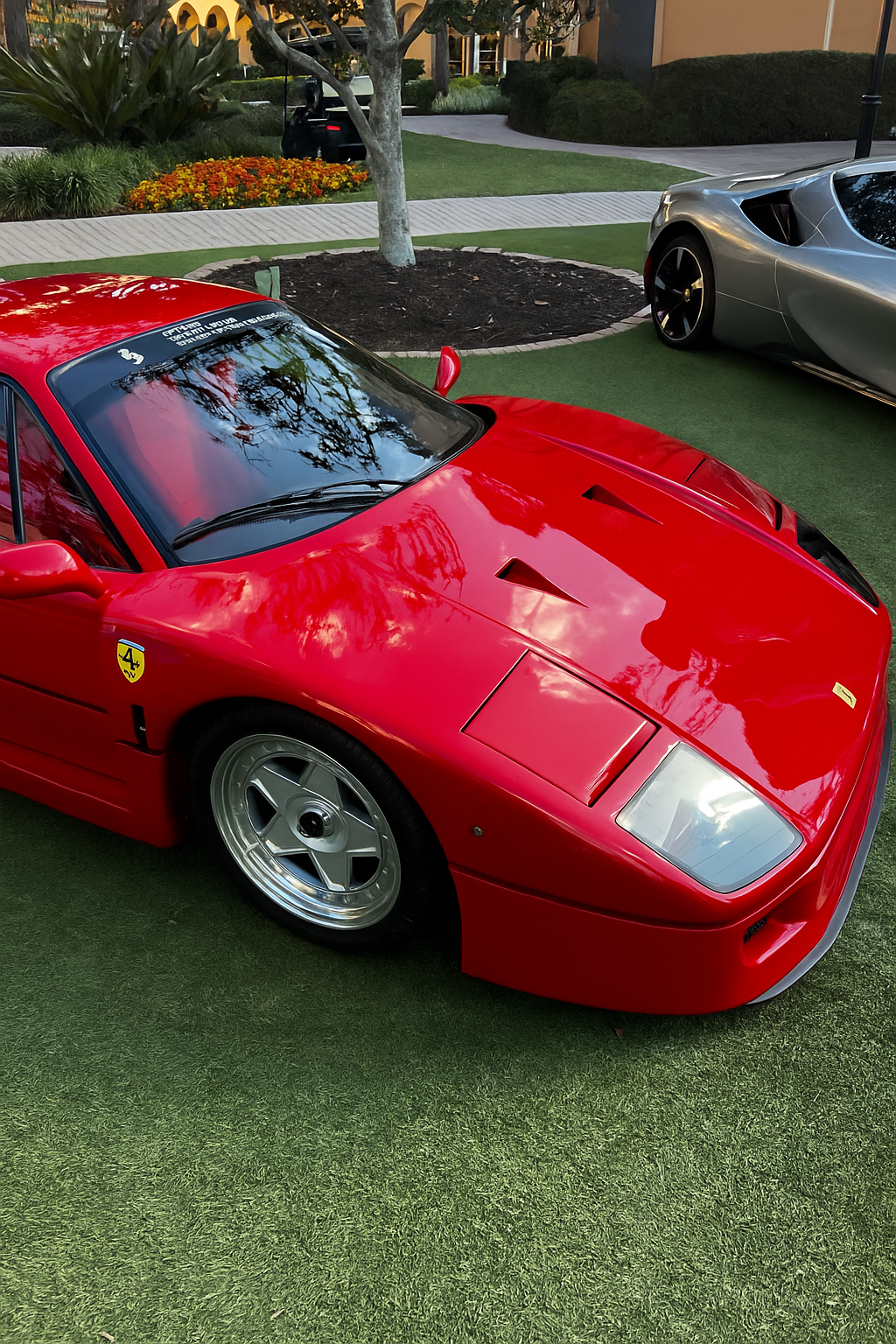 A vibrant red Ferrari F40 parked on a manicured lawn at an upscale residence, with a silver supercar visible in the background — capturing the caliber of elite clientele and iconic vehicles served by Voiture Noire.