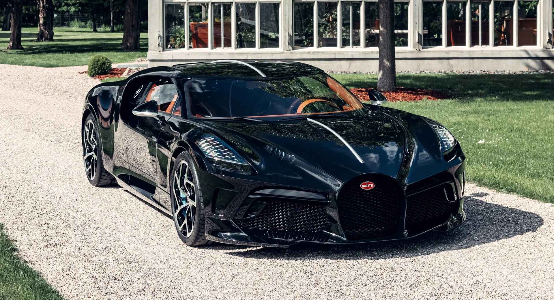 A sleek matte black Bugatti Voiture Noire parked on a gravel driveway, showcasing its dramatic low-profile body, sculpted carbon fiber panels, signature oval grille, and tan leather interior accents — embodying elite automotive craftsmanship.
