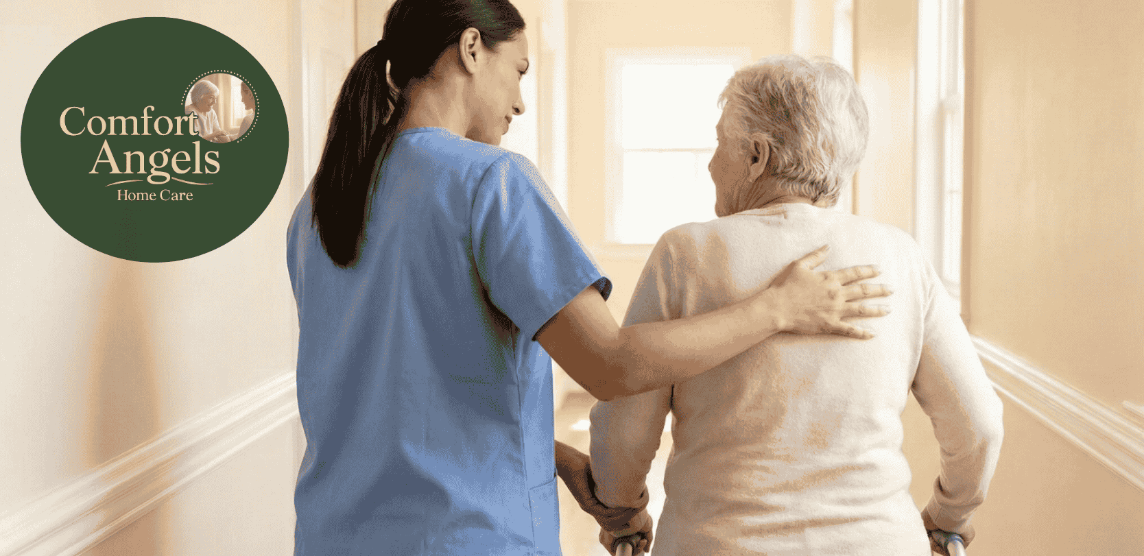 a caregiver in blue scrubs from behind, placing a supportive hand on the shoulder of an elderly woman who is using a walker.
