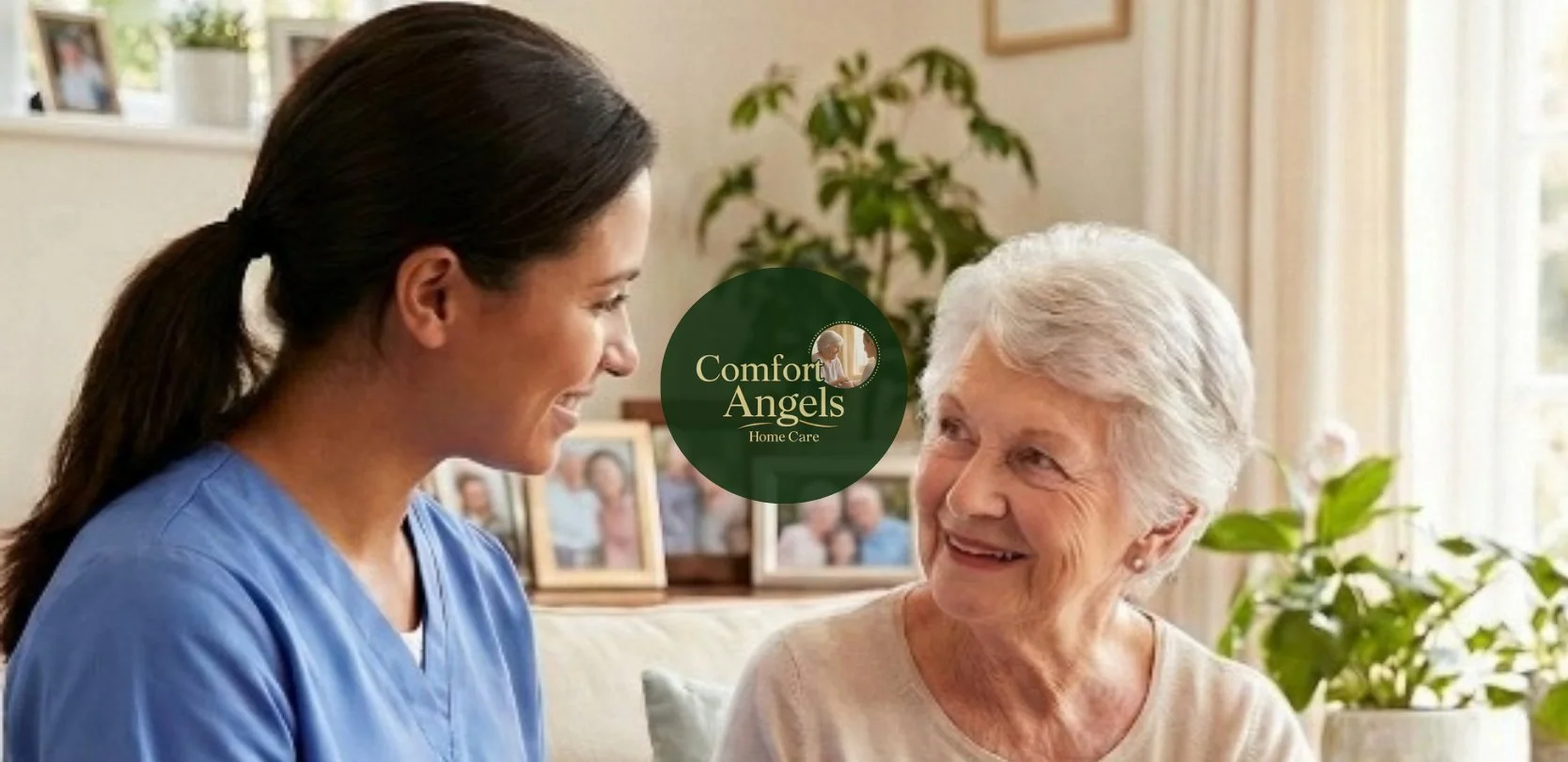 A young female caregiver in blue scrubs smiling and talking to an elderly woman with white hair, sitting in a well-lit living room, with green houseplants and picture frames in the background.