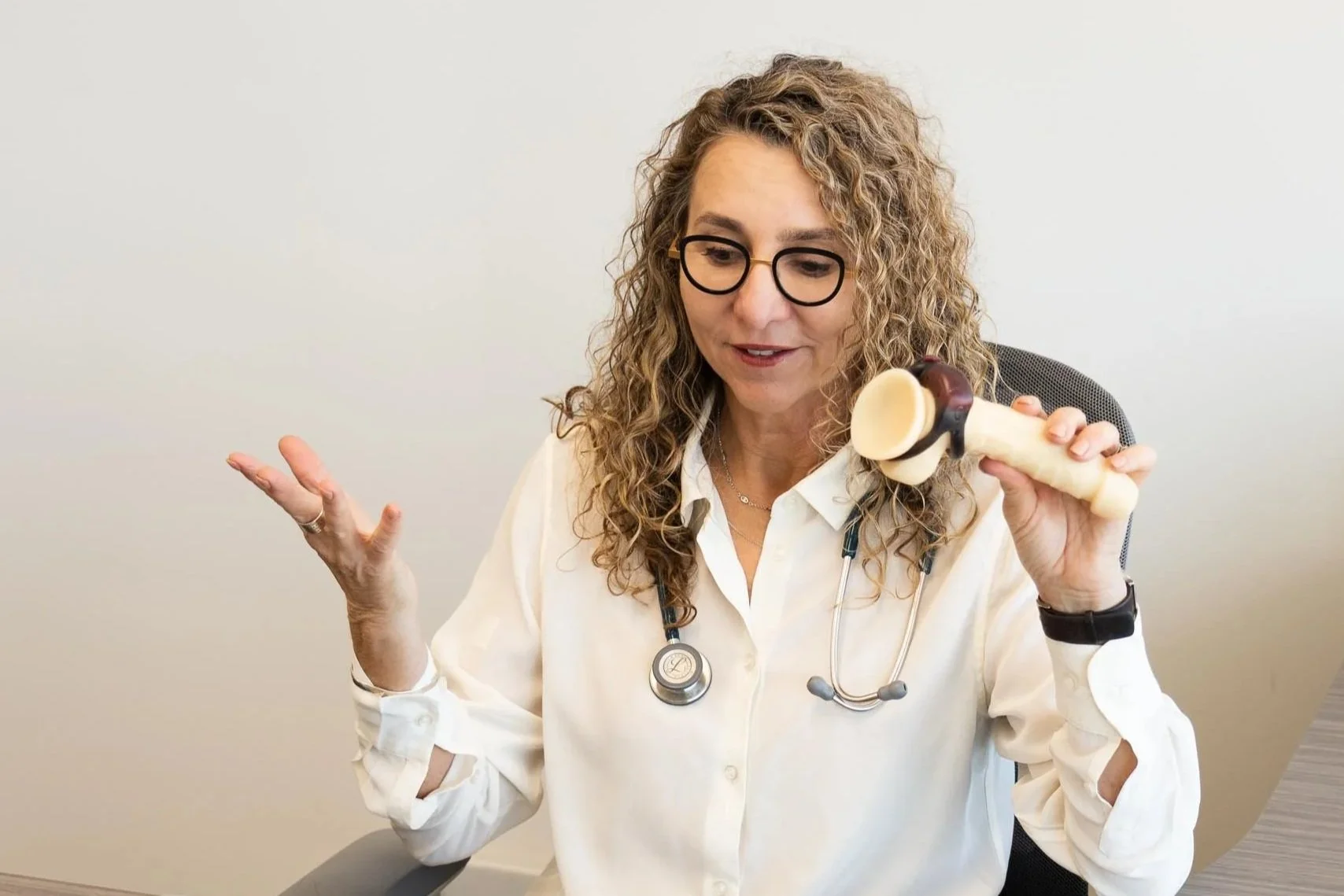 A female doctor with curly blonde hair, glasses, and a white lab coat, holding a large model of a human leg joint, sitting at a desk.