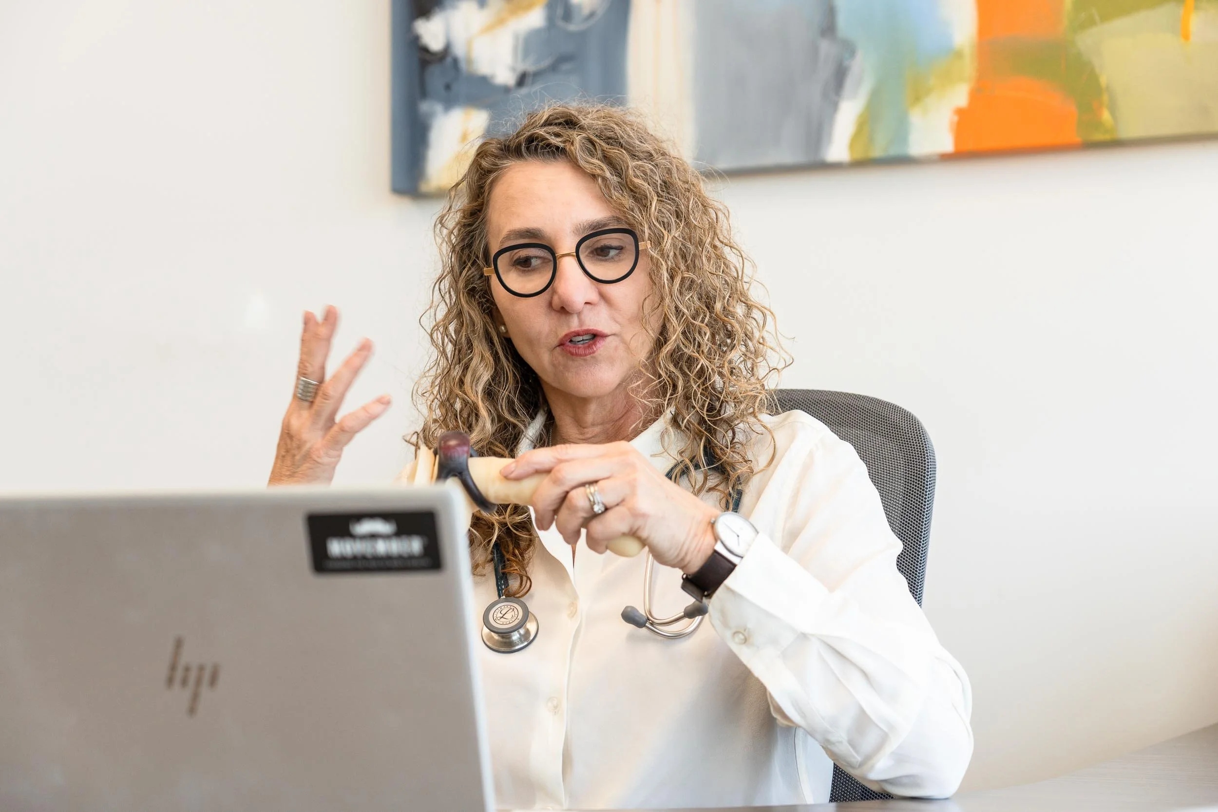 A woman wearing glasses and a white shirt sitting at a desk, holding a reflex hammer and a stethoscope, and talking in front of a silver laptop with a colorful abstract painting on the wall behind her.
