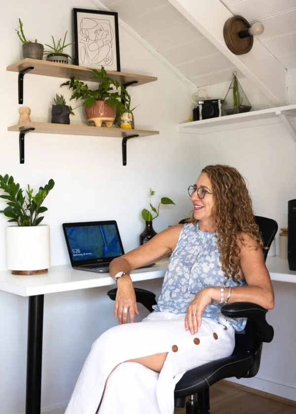 A woman with curly hair and glasses sitting in a black office chair at a white desk, smiling. The desk has a laptop, a potted plant, and a small plant in a black vase. Above, two wooden shelves hold various plants and decorative items, with framed art on the top shelf. The room has white walls and an angled ceiling with a exposed light bulb fixture.