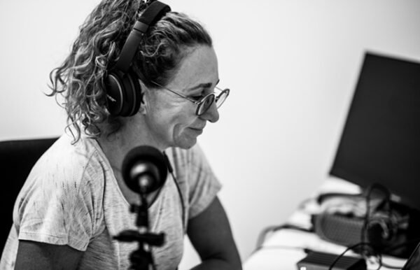 A woman wearing headphones and glasses working at a desk with a computer monitor and recording equipment.