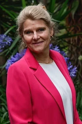 A woman with gray hair wearing a pink blazer and white top, standing outdoors in front of green foliage and purple flowers.
