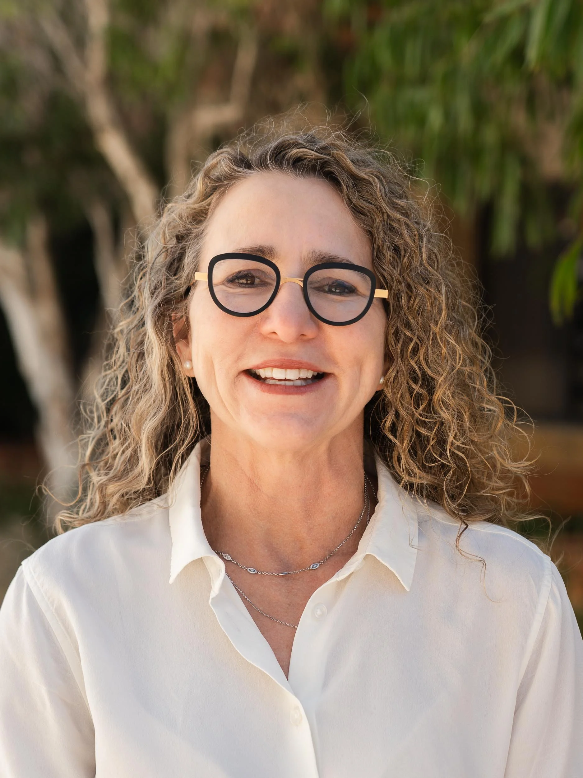 Close-up photo of a woman with curly blonde hair, wearing glasses, earrings, necklaces, and a white shirt, outdoors with trees in the background.