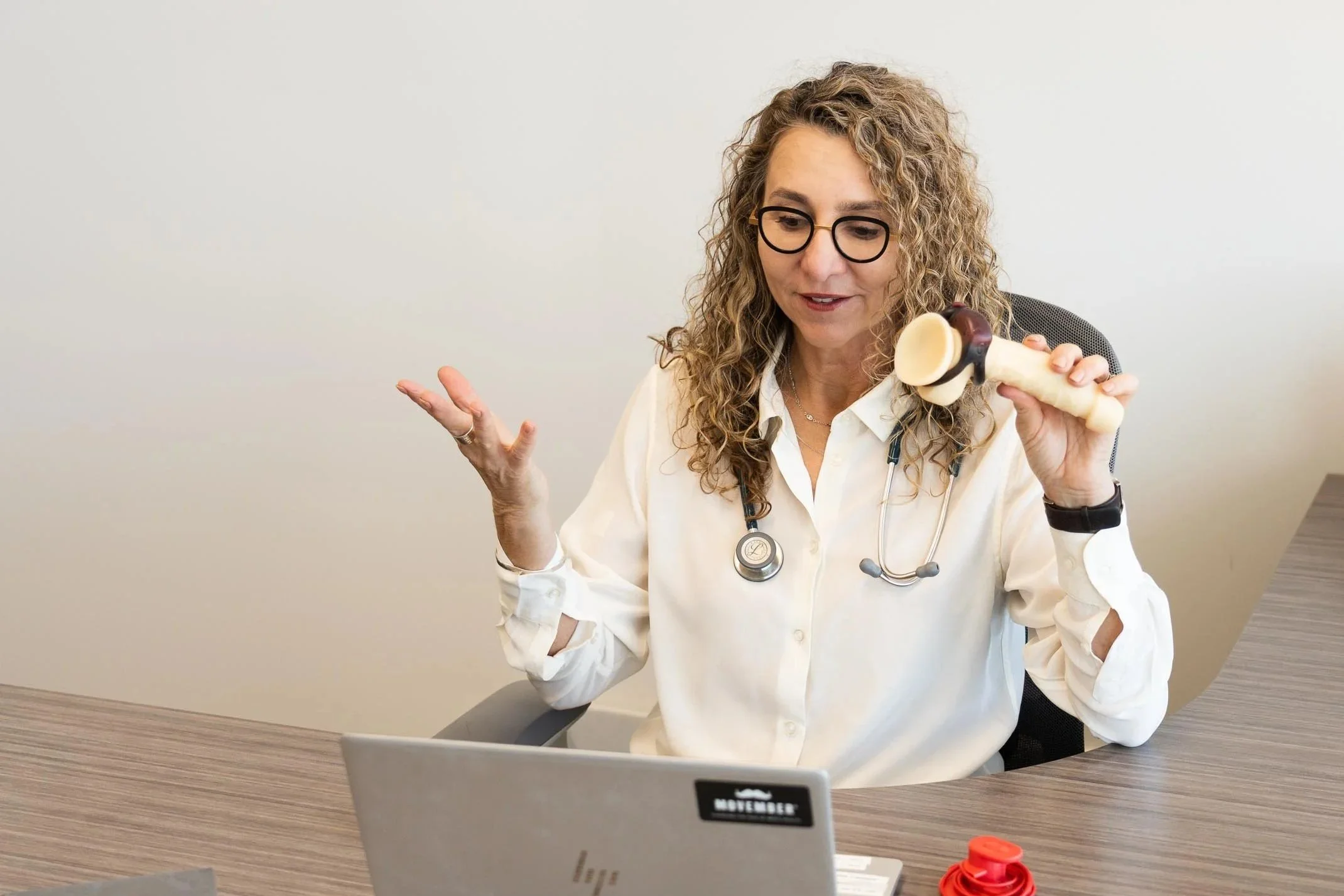 A woman with curly hair, glasses, and a stethoscope around her neck sitting at a desk, holding a bone-shaped object in one hand and gesturing with the other, with a laptop and a red object on the desk.