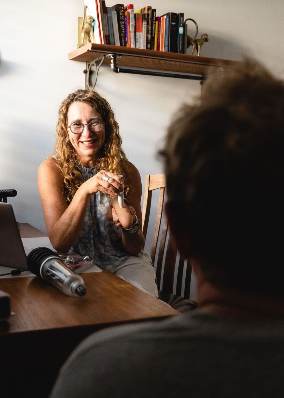 A woman with curly red hair, glasses, and a sleeveless patterned top sitting at a wooden table, smiling and engaging with a person whose back is to the camera. A bookshelf with books and decorative items is mounted on the wall behind her.