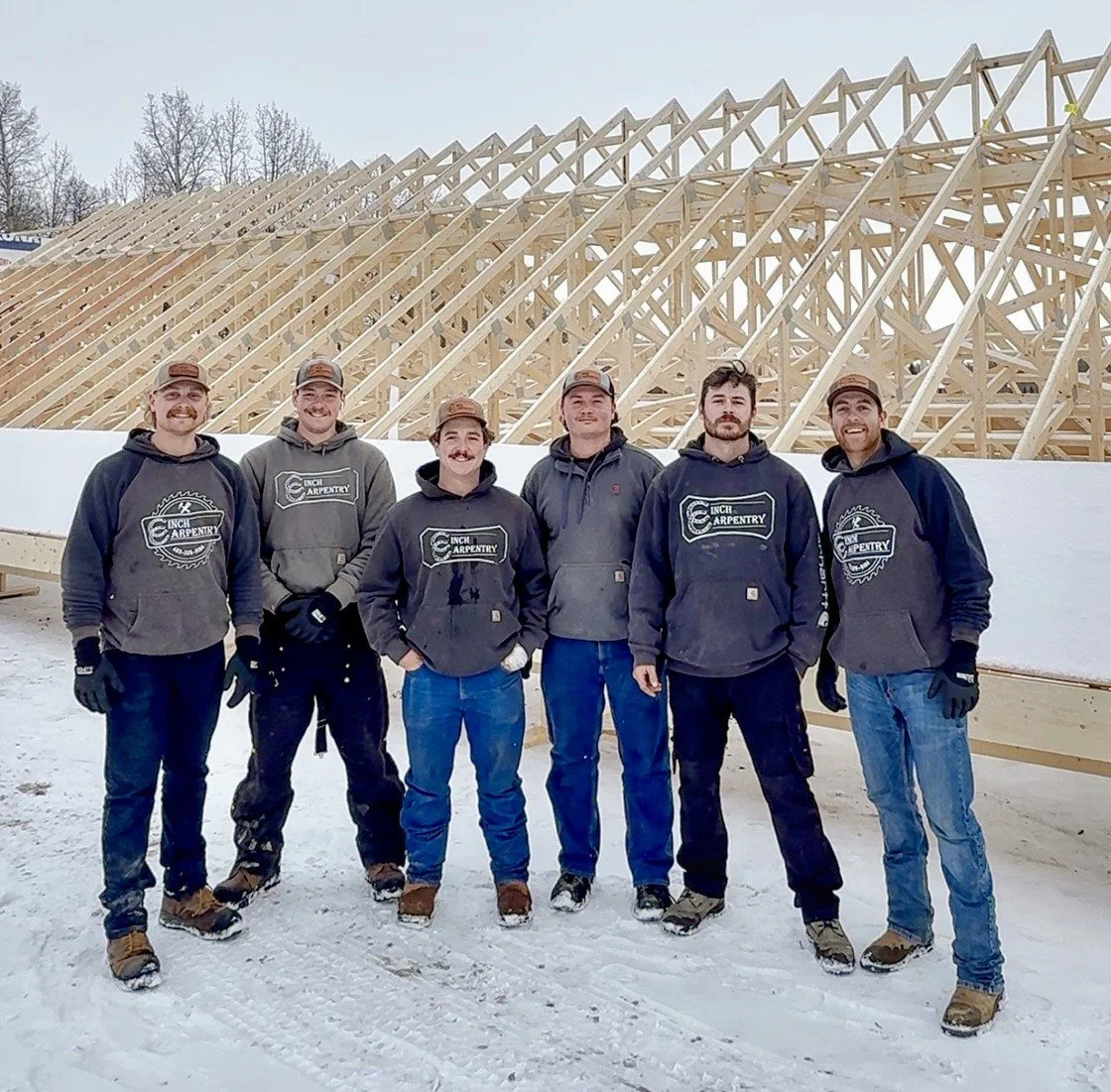 Six men standing in front of a large wooden building structure under construction in snowy weather, all wearing work clothes and hats.
