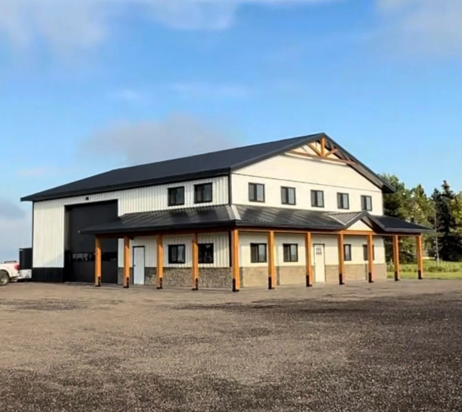 Aerial view of a construction site with wooden framing for a new building, adjacent to a parking lot with parked cars and a large blue industrial building.