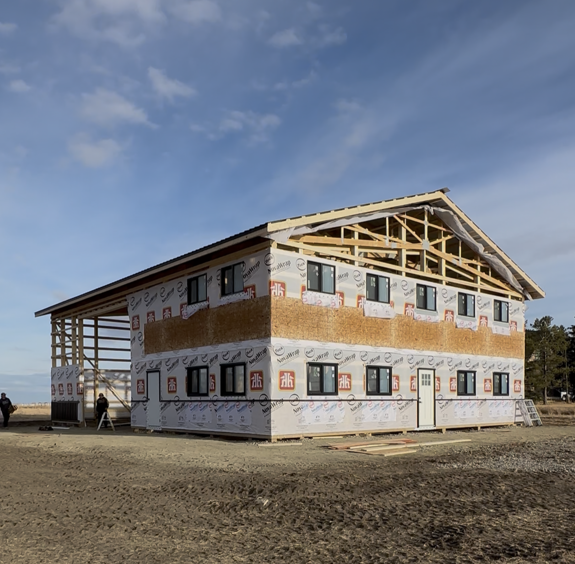 Newly constructed house with black siding and wooden support beams, situated on a dirt terrain surrounded by trees, under a blue sky with a few clouds.