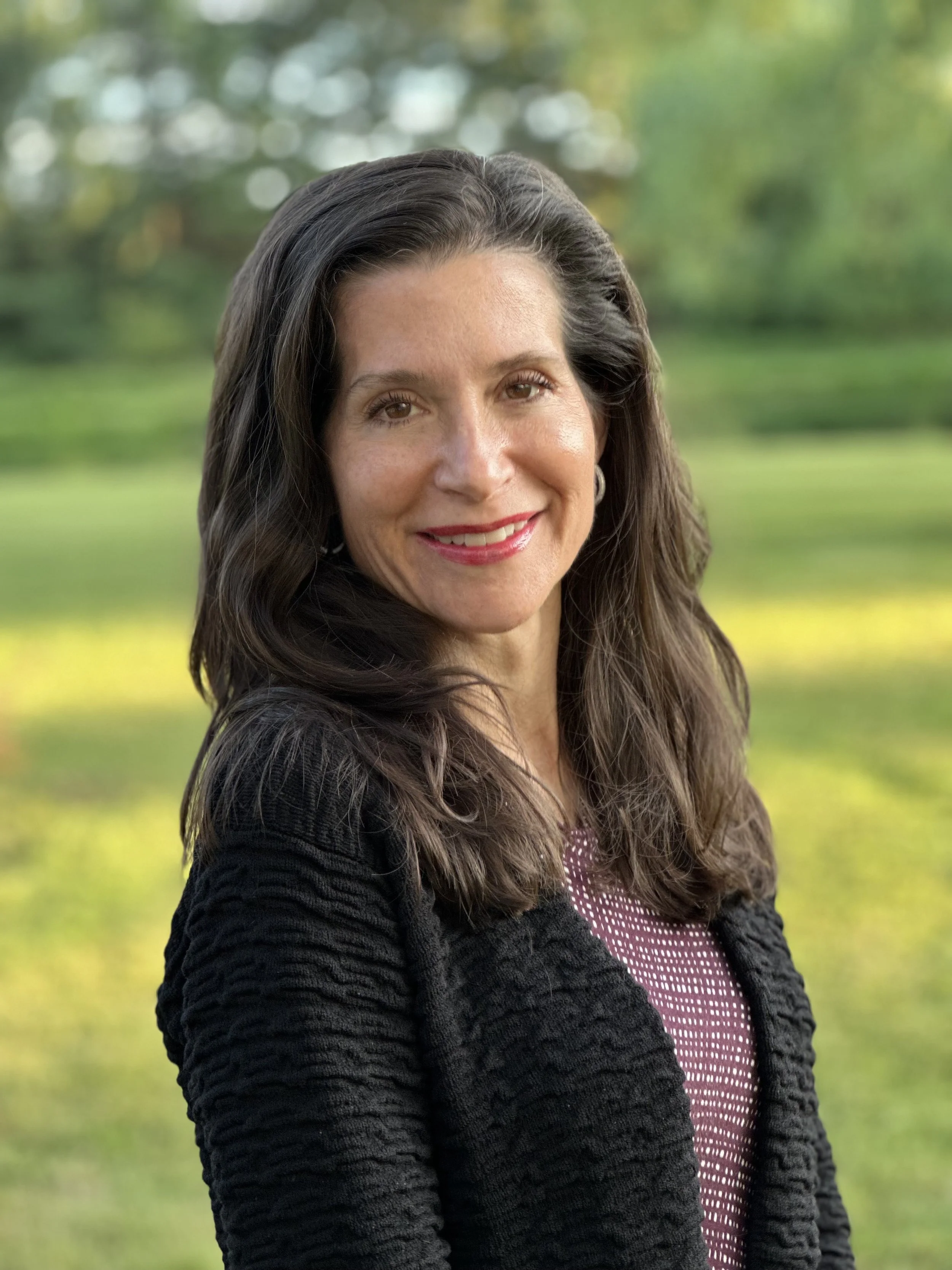 A woman with long dark hair and light skin, smiling outdoors with greenery in the background.