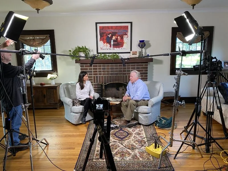 Interview set in a living room with two people seated on a sofa, surrounded by professional lighting and cameras.