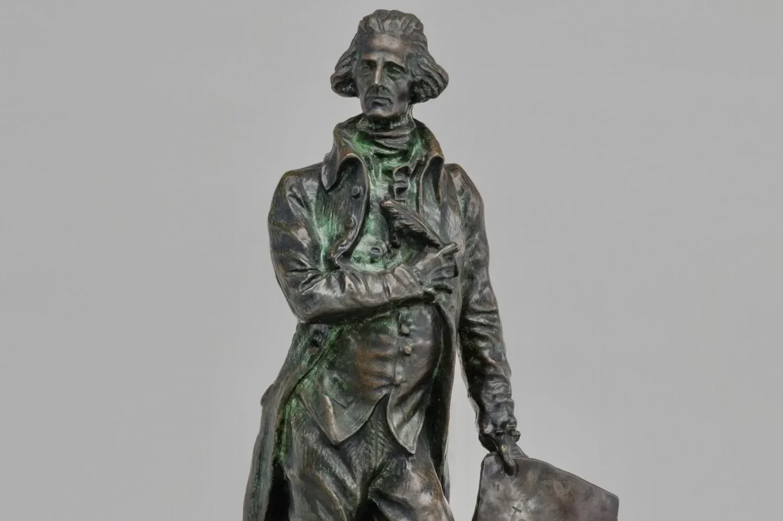 A bronze statue of a man dressed in 18th-century attire, holding a book and a quill, with a serious expression, set against a plain gray background.