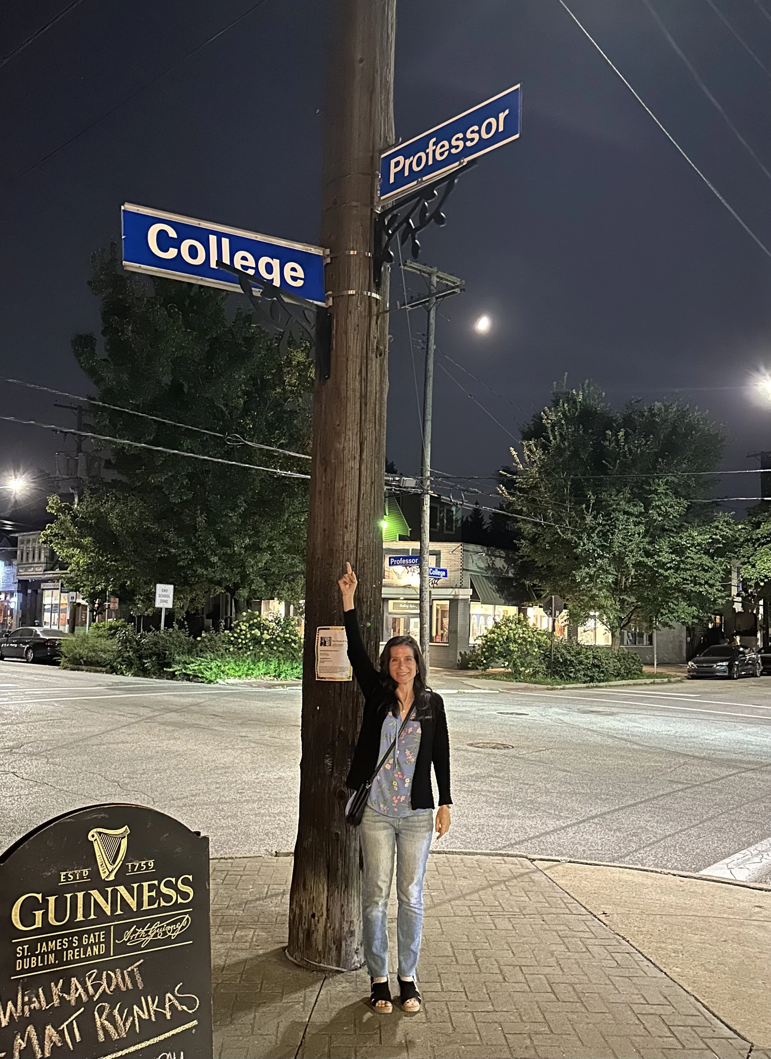 A woman standing on a city sidewalk at night, smiling and pointing upwards. She is next to a wooden utility pole with street signs labeled 'College' and 'Professor'. In the background, there are shops, trees, cars, streetlights, and a visible moon in the night sky. There is also a Guinness beer sign advertising a pub in Dublin, Ireland.