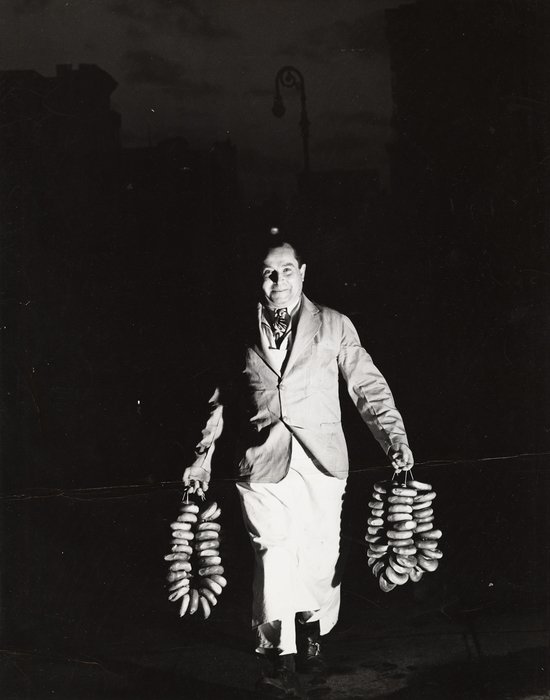 A person dressed in a suit walking at night carrying two strings of bagels, one in each hand.