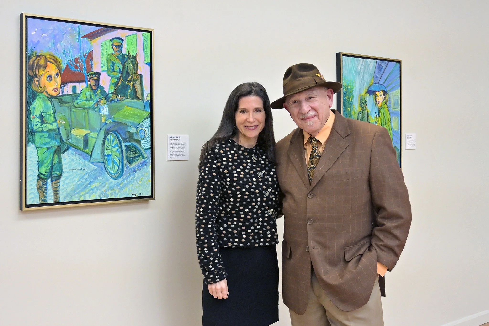 Samantha Baskind and Archie Rand standing in an art gallery, one woman and one man, with colorful paintings on the wall behind them.