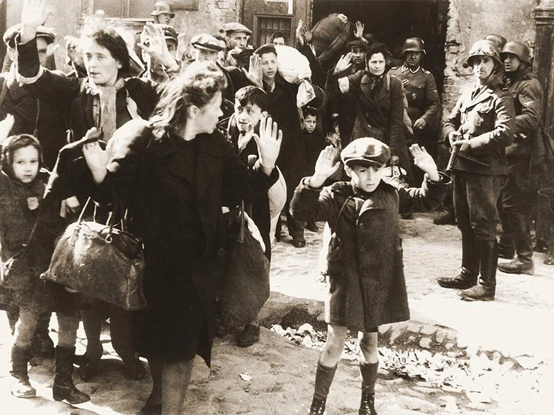 Women and children, some with backpacks, raising their hands as soldiers in helmets and military uniforms stand nearby, likely in a wartime or refugee scene.