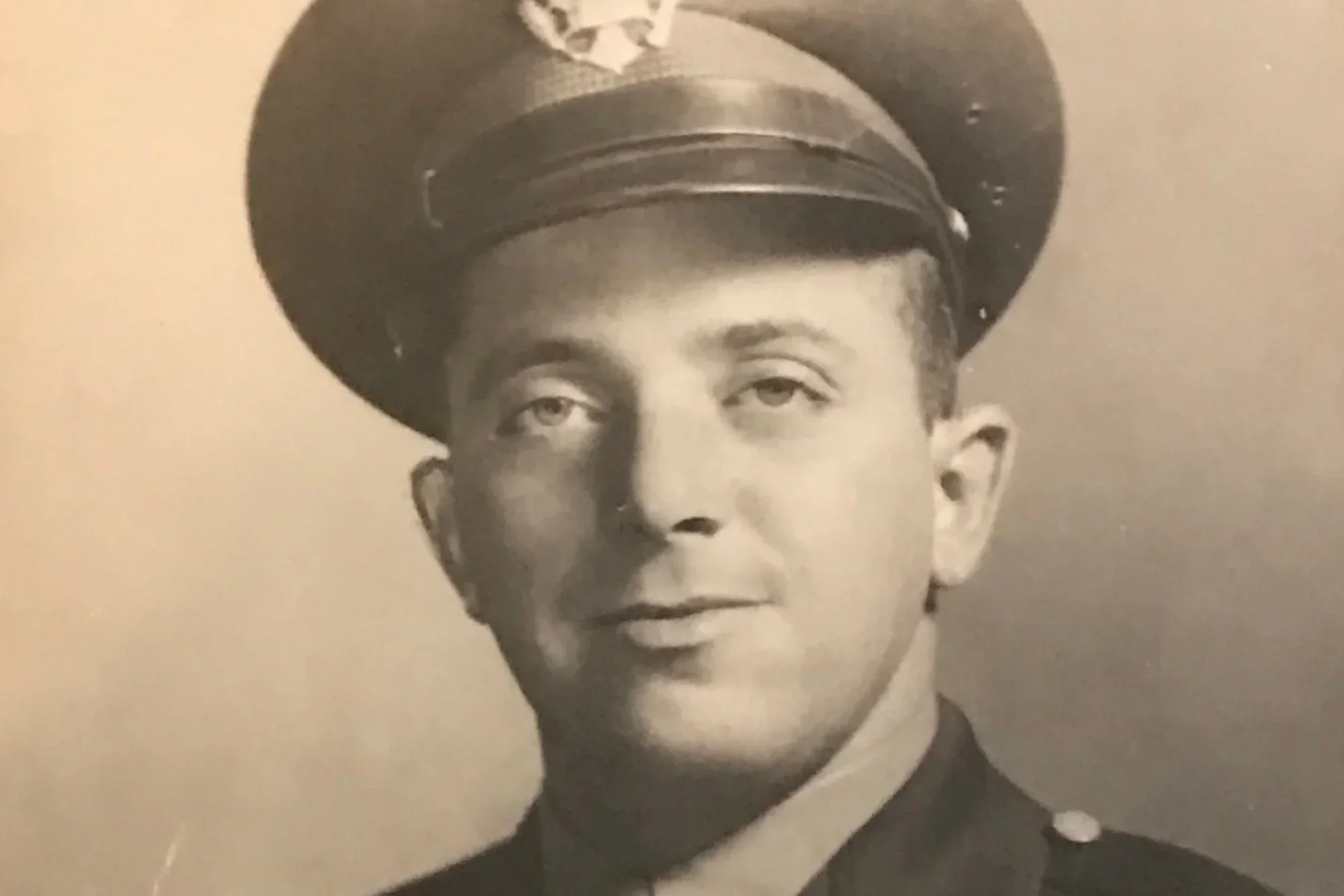 Black and white portrait of a young man in a military uniform, wearing a cap with a badge, looking at the camera with a slight smile.