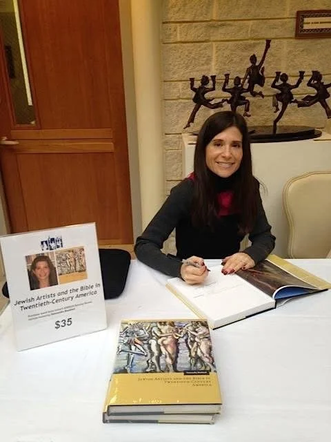 Samantha Baskind sitting at a table signing a book at a book signing event, with books and a sign displaying the book title and price on the table.