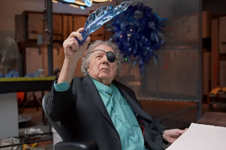 An elderly woman with an eye patch on her right eye, wearing a suit and light blue shirt, holding a glass sculpture up in her right hand, sitting in a workshop or studio with artistic materials and blue decorations in the background.