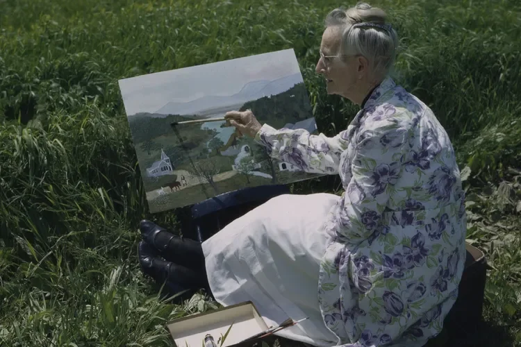 An elderly woman with short gray hair, wearing glasses and a floral shirt, sits across from the camera in tall grass. She is painting a landscape on a canvas that depicts a rural scene with mountains, water, and houses.