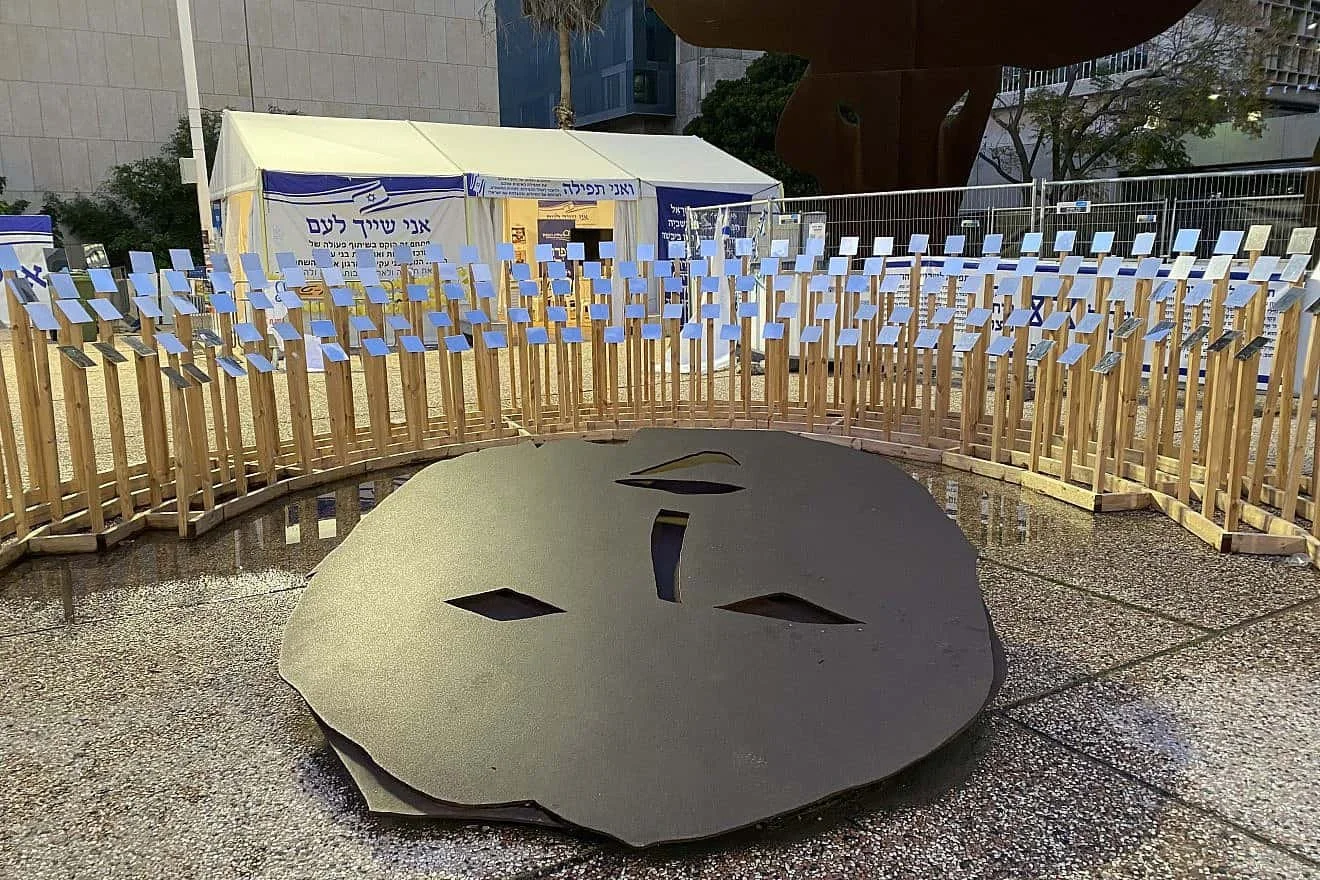 Outdoor art installation with a large black face mask sculpture on the ground in front of a tent with Hebrew writing and numerous small flags on a fence. Urban setting with buildings and trees.