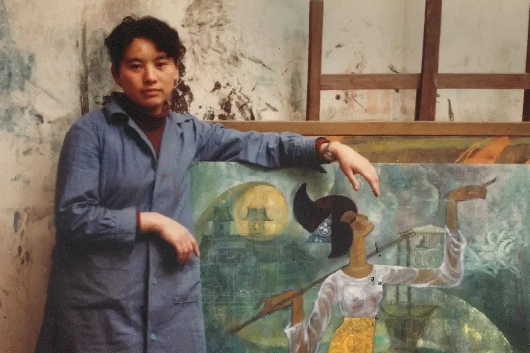 A woman with short dark hair wearing a blue jacket and red shirt pointing at a colorful mural painting of a woman with black hair, wearing a white blouse and yellow skirt, holding a spear, with a mystical landscape including a big moon, an elephant, a castle, and a mountain in the background.