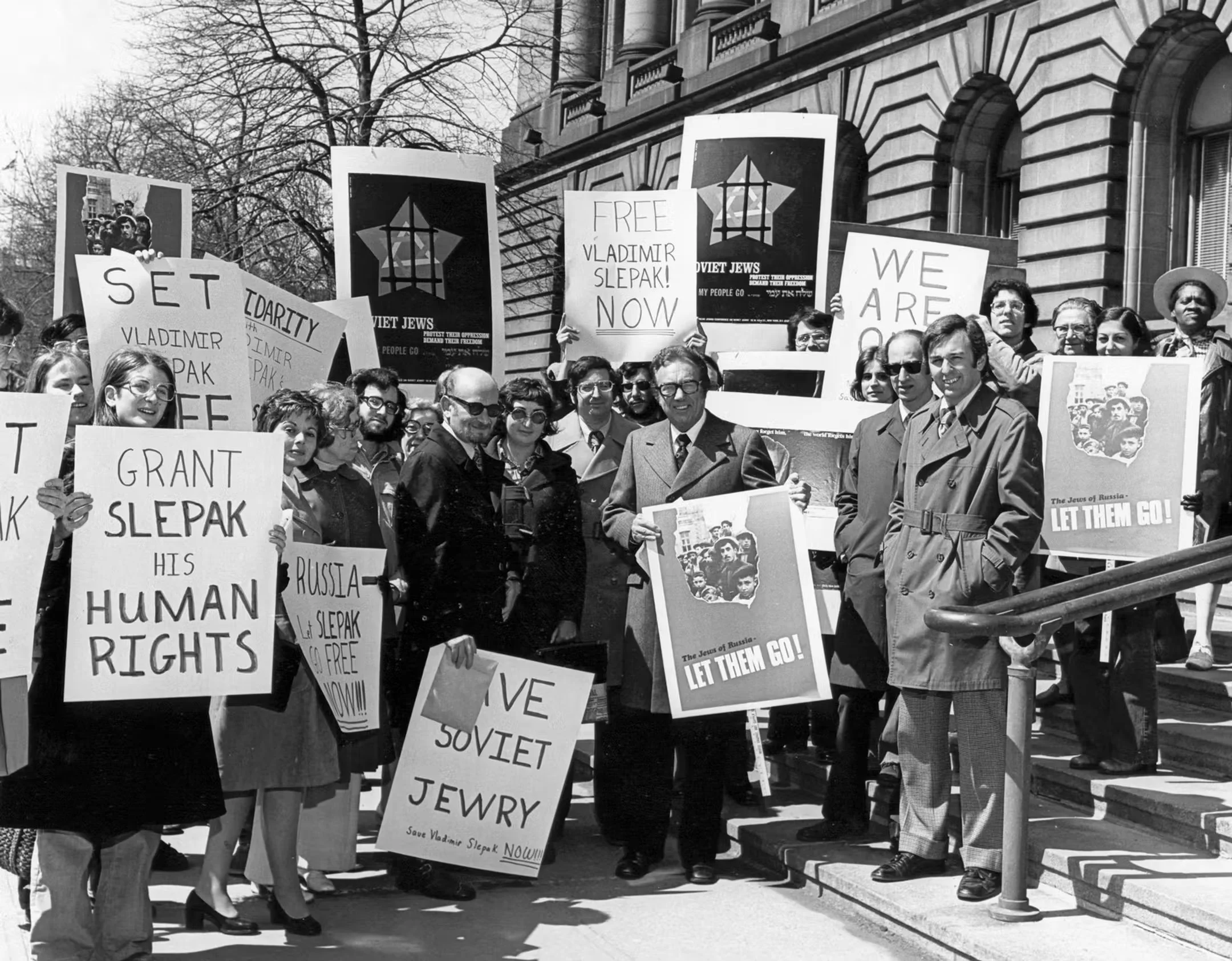 A black-and-white photo of a protest on the steps of a building with a crowd holding signs advocating for the release of Vladimir Slepak and supporting Jewish rights in Russia.