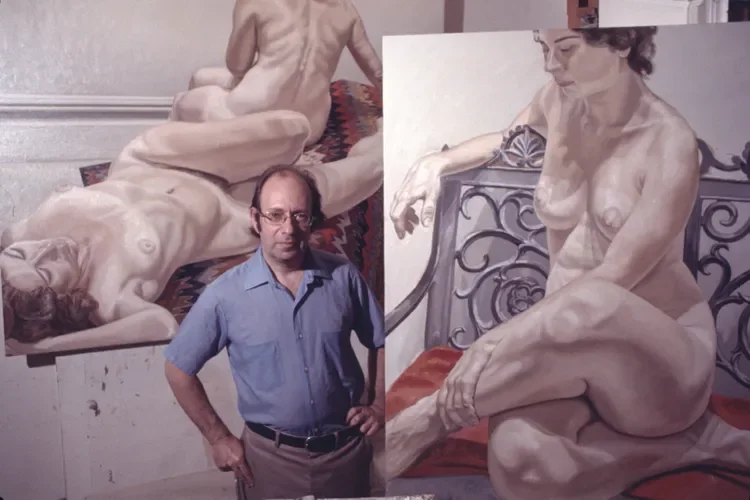 An artist standing in front of two large nude figure paintings, one of a reclining woman and one of a woman sitting, both in an art studio.