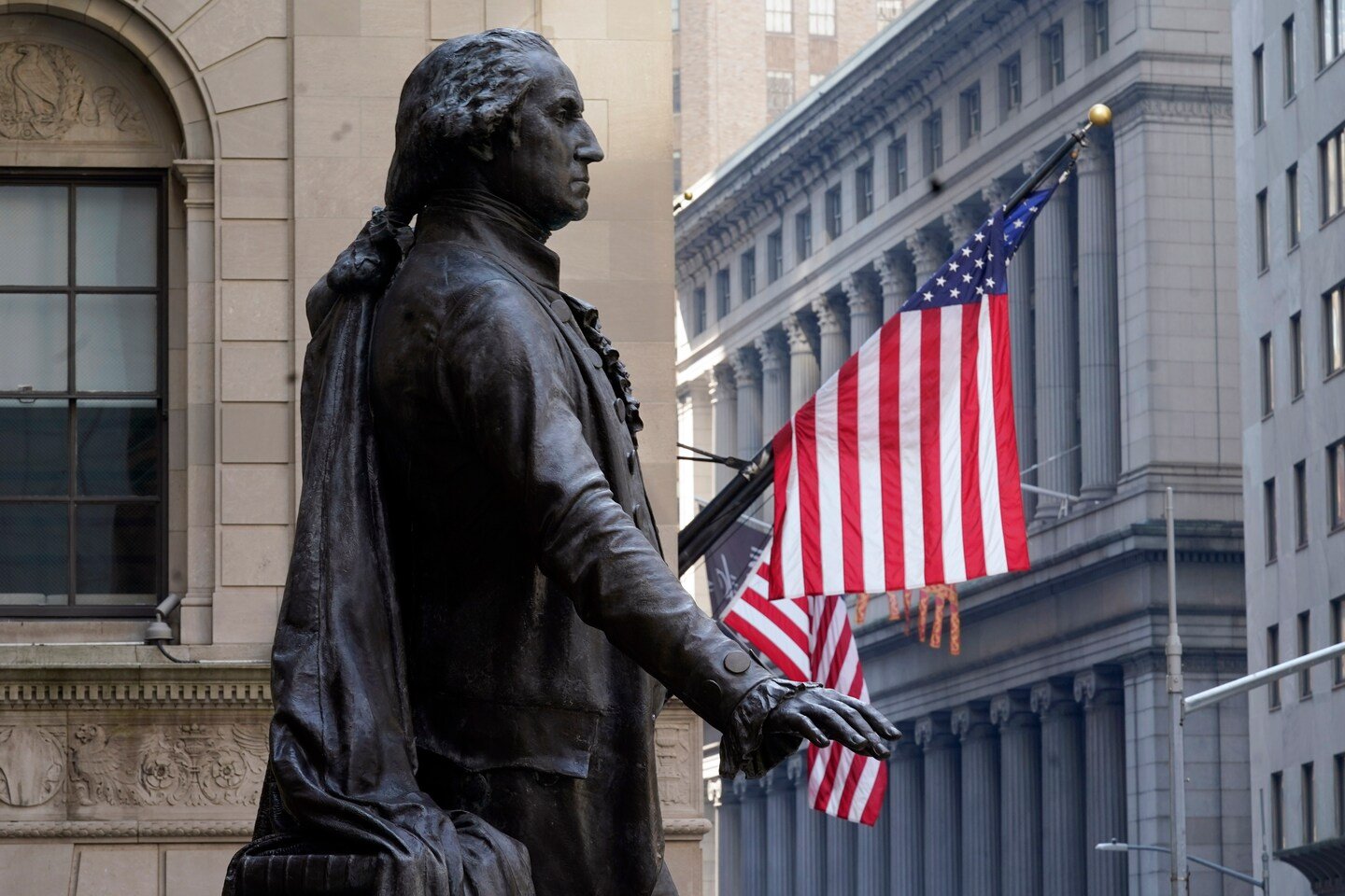 Bronze statue of a man in period clothing with long hair tied back, set against a city street background with American flags and tall buildings.