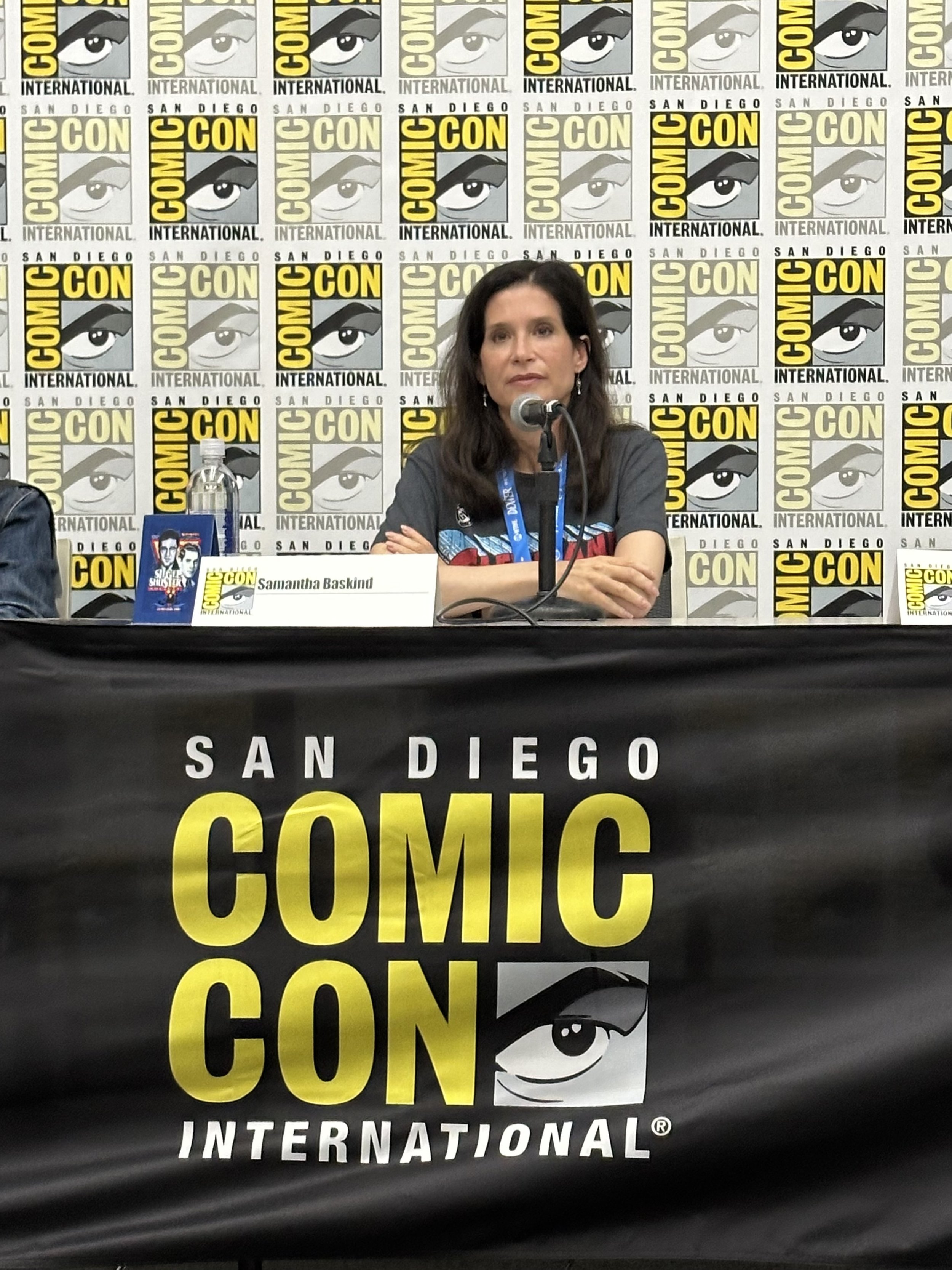 A woman, Samantha Baskind, seated at a panel table with a microphone, at San Diego Comic-Con International, with a backdrop of Comic-Con logos. The table banner features the Comic-Con logo.