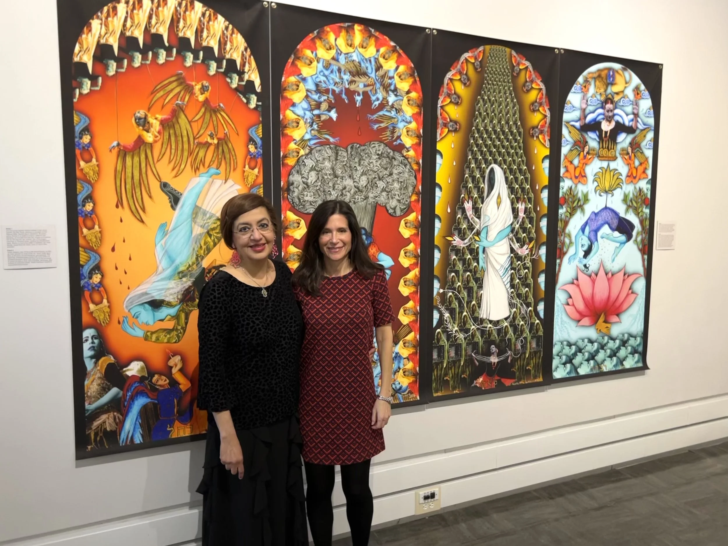 Samantha Baskind and Siona Benjamin standing in front of colorful, detailed artwork in an art gallery. The artwork includes vivid imagery and stylized figures with vibrant backgrounds.