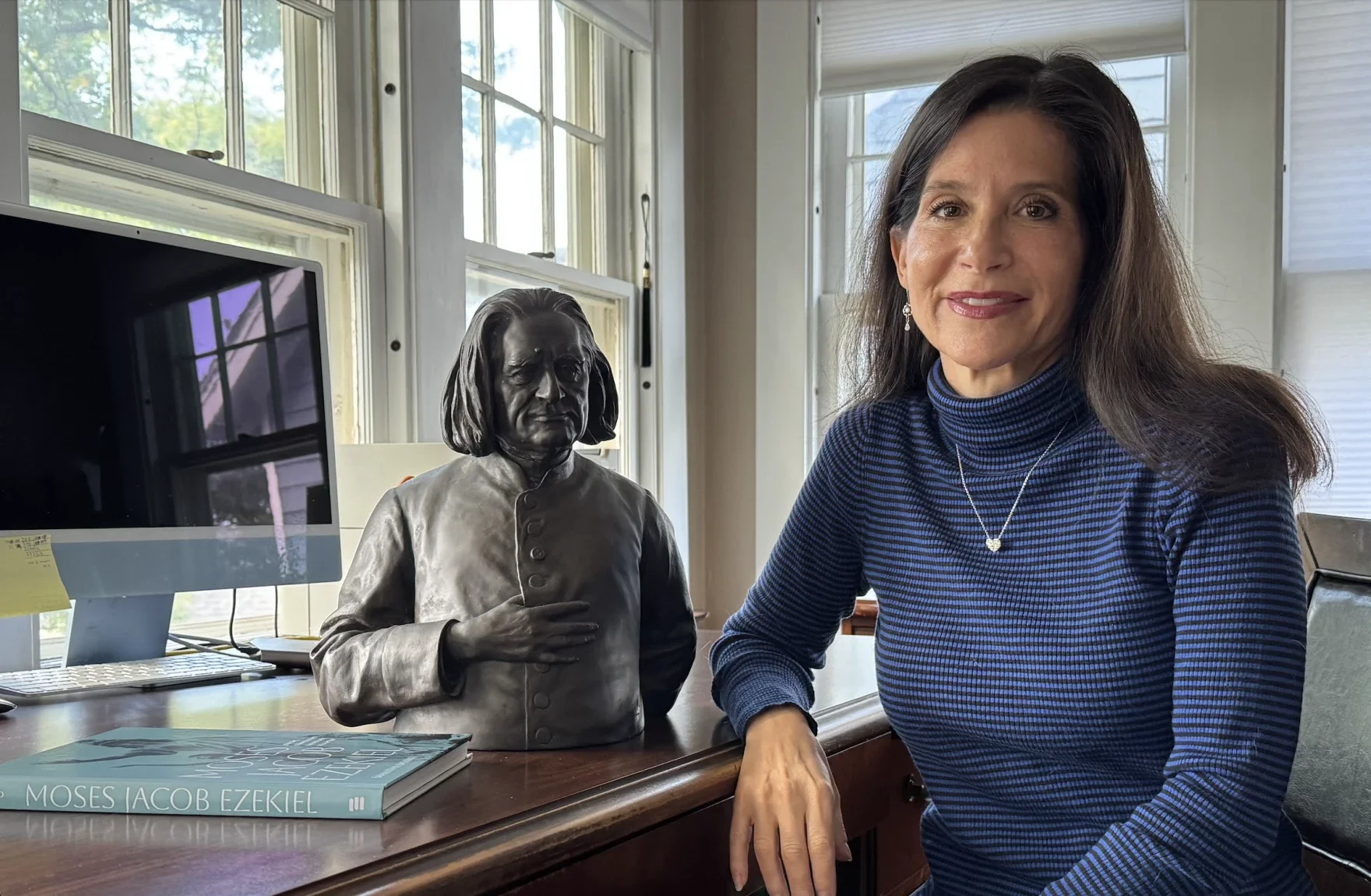 Samantha Baskind sitting next to a bronze bust of a woman, on a wooden desk in a sunlit room with large windows.